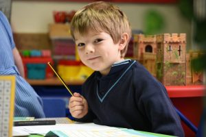Reception pupil sat at desk with pencil and book smiling